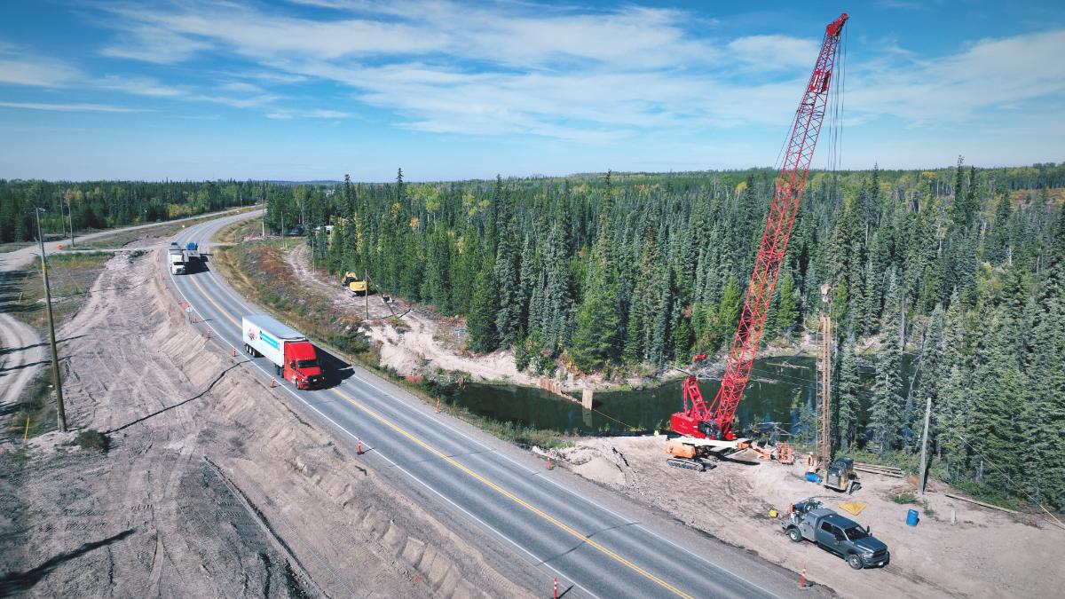 Aerial view of a crawler crane driving pile beside a live two-lane highway